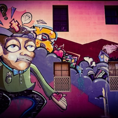 A colorful street art mural featuring a cartoonish character with an exaggerated facial expression, holding a heart in its hand. Surrounding the character are abstract shapes, including clouds, a speech bubble with a diamond, and various urban elements like a house and a can. The background includes a building wall with windows.