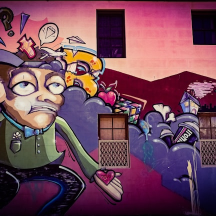 A colorful street art mural featuring a cartoonish character with an exaggerated facial expression, holding a heart in its hand. Surrounding the character are abstract shapes, including clouds, a speech bubble with a diamond, and various urban elements like a house and a can. The background includes a building wall with windows.