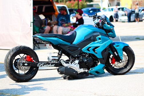 A custom-designed blue motorcycle is parked in a parking lot. The bike features a striking design with angular panels and details in black and red. A group of people is blurred in the background, sitting and standing near cars and a canopy tent.