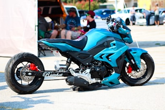 A custom-designed blue motorcycle is parked in a parking lot. The bike features a striking design with angular panels and details in black and red. A group of people is blurred in the background, sitting and standing near cars and a canopy tent.