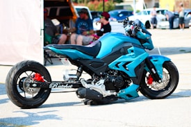 A custom-designed blue motorcycle is parked in a parking lot. The bike features a striking design with angular panels and details in black and red. A group of people is blurred in the background, sitting and standing near cars and a canopy tent.