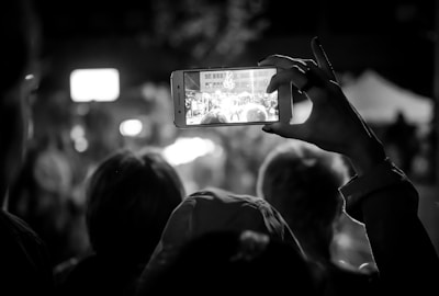 In a dimly lit setting, a person holds up a smartphone, capturing a bustling scene in front of them. The phone's screen displays a clearer view of a gathering, possibly a crowd at an event. The outlines of several heads can be seen, slightly out of focus, indicating an audience or spectators. The composition focuses on the act of capturing a moment digitally within a live experience.