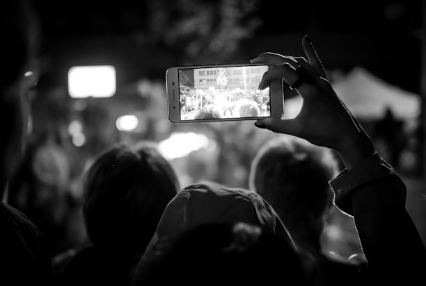 In a dimly lit setting, a person holds up a smartphone, capturing a bustling scene in front of them. The phone's screen displays a clearer view of a gathering, possibly a crowd at an event. The outlines of several heads can be seen, slightly out of focus, indicating an audience or spectators. The composition focuses on the act of capturing a moment digitally within a live experience.