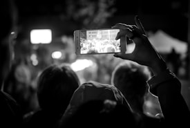 In a dimly lit setting, a person holds up a smartphone, capturing a bustling scene in front of them. The phone's screen displays a clearer view of a gathering, possibly a crowd at an event. The outlines of several heads can be seen, slightly out of focus, indicating an audience or spectators. The composition focuses on the act of capturing a moment digitally within a live experience.