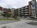 A modern apartment complex with multiple floors, characterized by geometric patterns and a colorful mural on the side of the building. The structure is surrounded by a small parking lot with several parked cars and some greenery including neatly aligned small trees and bushes.