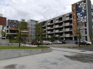 A modern apartment complex with multiple floors, characterized by geometric patterns and a colorful mural on the side of the building. The structure is surrounded by a small parking lot with several parked cars and some greenery including neatly aligned small trees and bushes.