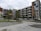 A modern apartment complex with multiple floors, characterized by geometric patterns and a colorful mural on the side of the building. The structure is surrounded by a small parking lot with several parked cars and some greenery including neatly aligned small trees and bushes.