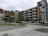 A modern apartment complex with multiple floors, characterized by geometric patterns and a colorful mural on the side of the building. The structure is surrounded by a small parking lot with several parked cars and some greenery including neatly aligned small trees and bushes.