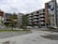 A modern apartment complex with multiple floors, characterized by geometric patterns and a colorful mural on the side of the building. The structure is surrounded by a small parking lot with several parked cars and some greenery including neatly aligned small trees and bushes.