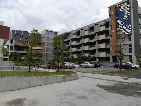 A modern apartment complex with multiple floors, characterized by geometric patterns and a colorful mural on the side of the building. The structure is surrounded by a small parking lot with several parked cars and some greenery including neatly aligned small trees and bushes.