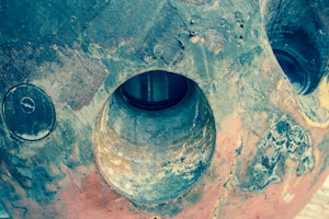An aged, metallic surface with circular openings and a textured, weathered appearance. The colors range from dark and rusty brown to faded blue-green. The surface shows signs of corrosion and wear, with patches of discoloration.