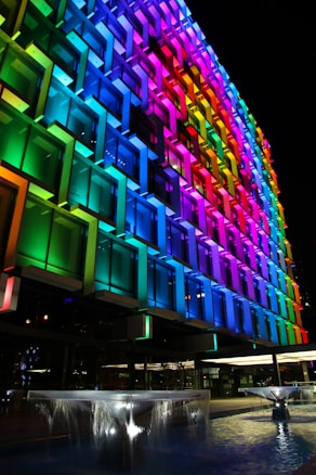 A building facade illuminated with a vibrant spectrum of colors ranging from red to violet. The design features a geometric pattern with protruding square panels, each highlighted by colorful lights. In the foreground, there are fountains with cascading water, lit from below, adding a dynamic element to the scene.