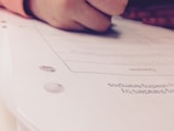 A close-up view of a hand holding a pen while writing or marking on a sheet of paper. The focus is primarily on the action of writing, with the text and details on the paper being slightly out of focus.