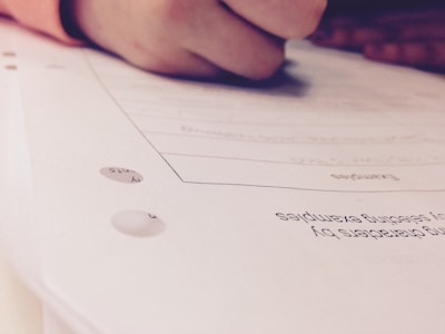 A close-up view of a hand holding a pen while writing or marking on a sheet of paper. The focus is primarily on the action of writing, with the text and details on the paper being slightly out of focus.