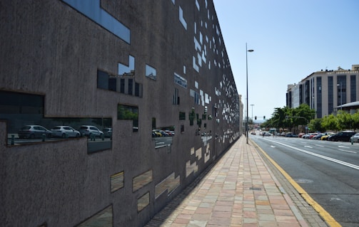 A modern architectural wall featuring numerous rectangular reflective glass panels arranged in an irregular pattern. The wall runs parallel to a street with visible cars parked alongside it. Buildings with contemporary designs can be seen in the background, interspersed with lush green trees. The pavement is made of variously colored rectangular tiles, and the sky is clear and sunny.