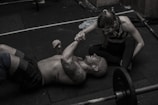 A man is lying on a gym floor with a woman kneeling beside him, holding his hand. Both appear to be in athletic attire. There are weights and gym equipment scattered around them, suggesting a workout session. The man has tattoos on his arm and is smiling at the woman, conveying a sense of camaraderie or encouragement.