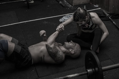 A man is lying on a gym floor with a woman kneeling beside him, holding his hand. Both appear to be in athletic attire. There are weights and gym equipment scattered around them, suggesting a workout session. The man has tattoos on his arm and is smiling at the woman, conveying a sense of camaraderie or encouragement.