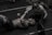 A man is lying on a gym floor with a woman kneeling beside him, holding his hand. Both appear to be in athletic attire. There are weights and gym equipment scattered around them, suggesting a workout session. The man has tattoos on his arm and is smiling at the woman, conveying a sense of camaraderie or encouragement.