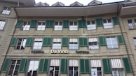 The building features a traditional architectural style with multiple windows equipped with green shutters. The signage indicates a pizzeria and pasticceria named 'Da Vinci'. The facade is beige, and there are several awnings above the windows, some of which are striped.