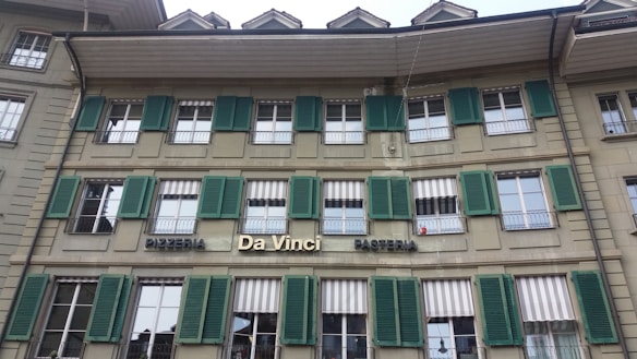 The building features a traditional architectural style with multiple windows equipped with green shutters. The signage indicates a pizzeria and pasticceria named 'Da Vinci'. The facade is beige, and there are several awnings above the windows, some of which are striped.