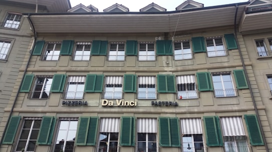 The building features a traditional architectural style with multiple windows equipped with green shutters. The signage indicates a pizzeria and pasticceria named 'Da Vinci'. The facade is beige, and there are several awnings above the windows, some of which are striped.