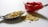 A vivid red chili pepper is placed on a white surface next to a metallic spoon filled with yellow seeds. Some of the seeds are scattered on the surface beside the spoon, creating a textured and colorful composition.