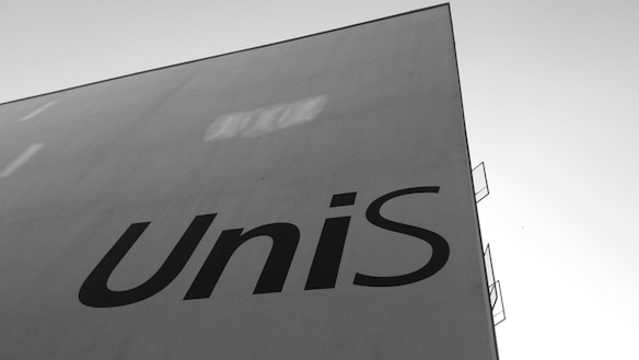 A large, flat, white building facade with the word 'UniS' in bold, black letters. The sky is clear and the perspective is from a low angle, emphasizing the height and simplicity of the structure.