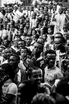 A large group of people gathered together, predominantly smiling and appearing engaged. The crowd includes men, women, and children of various ages. The focus seems to be on the interaction and expressions among the people, with some individuals clapping and others raising their hands.
