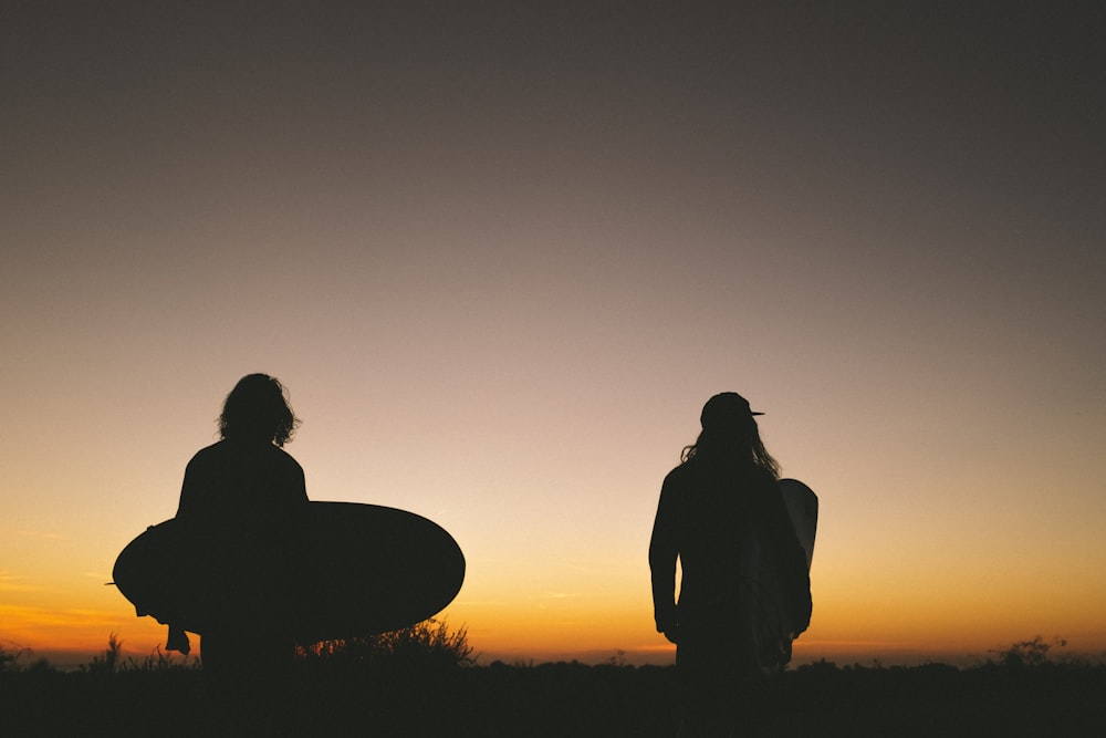 two person walking on road while holding surfboard during golden hour