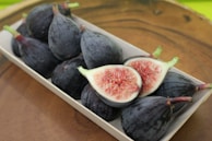 A wooden surface holds a rectangular tray filled with dark purple figs. Two figs are halved, revealing their pink, seedy interior.
