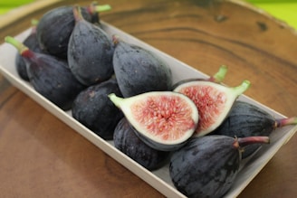 A wooden surface holds a rectangular tray filled with dark purple figs. Two figs are halved, revealing their pink, seedy interior.