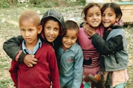 Five children of diverse appearances stand closely together, smiling warmly at the camera. They are dressed in various jackets and sweaters, showcasing a range of colors from red to pink to blue. Behind them is an outdoor setting with grass and rocks, suggesting a casual, natural environment.