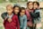 Five children of diverse appearances stand closely together, smiling warmly at the camera. They are dressed in various jackets and sweaters, showcasing a range of colors from red to pink to blue. Behind them is an outdoor setting with grass and rocks, suggesting a casual, natural environment.