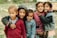 Five children of diverse appearances stand closely together, smiling warmly at the camera. They are dressed in various jackets and sweaters, showcasing a range of colors from red to pink to blue. Behind them is an outdoor setting with grass and rocks, suggesting a casual, natural environment.
