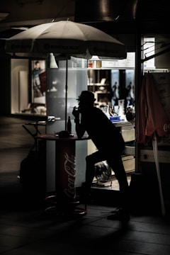 A silhouetted figure leans against a small outdoor kiosk under a large Coca-Cola branded umbrella. The scene is dimly lit, with the interior of the kiosk faintly illuminated. Various items and equipment are visible inside the kiosk, and there are blurry, indistinct figures in the background, suggesting an urban environment.
