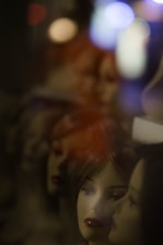 Multiple mannequin heads with realistic facial features are seen in varying focus. The background is dark, with spots of colorful blurred lights creating a dreamy or surreal atmosphere.