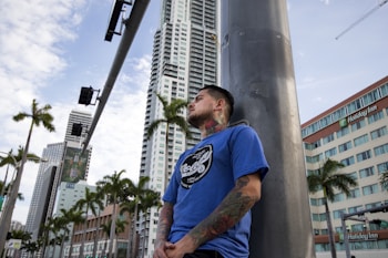 A tattooed person in a blue shirt leans against a large pole on a city street. Tall buildings, including a Holiday Inn, and palm trees are visible in the background, under a partly cloudy sky.