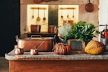 A rustic kitchen setting features copper pots and utensils hanging on the wall. The countertop displays a mix of decorative items including antique copper pots, lush green plants, and exotic fruits like a durian. The background exhibits a warm, cozy ambiance with a worn, vintage look.