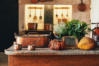 A rustic kitchen setting features copper pots and utensils hanging on the wall. The countertop displays a mix of decorative items including antique copper pots, lush green plants, and exotic fruits like a durian. The background exhibits a warm, cozy ambiance with a worn, vintage look.