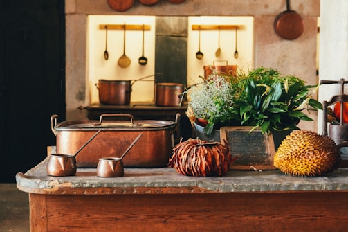 A rustic kitchen setting features copper pots and utensils hanging on the wall. The countertop displays a mix of decorative items including antique copper pots, lush green plants, and exotic fruits like a durian. The background exhibits a warm, cozy ambiance with a worn, vintage look.