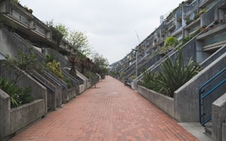 A modernist architectural scene featuring symmetrical concrete structures on both sides of a wide, red-brick pathway. The buildings have a series of staggered steps and balconies, each adorned with various green plants. The sky is overcast, adding a muted atmosphere to the overall setting.