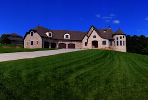 A large, elegant stone house with multiple gables and a turret sits atop a lush green hill under a vibrant blue sky. The house features several dormer windows and a spacious driveway leading up to it. Another house is visible in the background on the left.