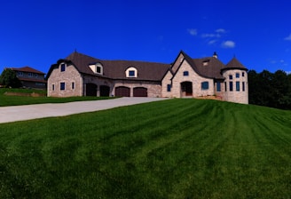A large, elegant stone house with multiple gables and a turret sits atop a lush green hill under a vibrant blue sky. The house features several dormer windows and a spacious driveway leading up to it. Another house is visible in the background on the left.
