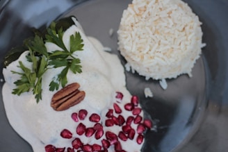 A gourmet dish consisting of a poblano pepper covered in a creamy white sauce, garnished with fresh parsley and a pecan, accompanied by scattered pomegranate seeds. A mound of white rice is served on the side.