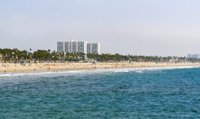 A sandy beach stretching along the coastline with people enjoying various activities on both the sand and in the water. Tall palm trees line the background, and there is a large modern building in the distance.
