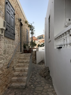 A narrow cobblestone alley flanked by stone and white-painted buildings, leading to a residential area with plants and staircases. The alley has a Mediterranean feel, with rustic charm and traditional architectural details.