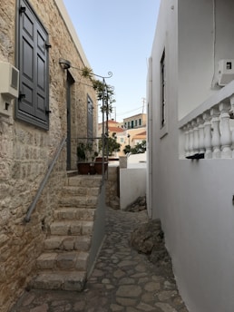 A narrow cobblestone alley flanked by stone and white-painted buildings, leading to a residential area with plants and staircases. The alley has a Mediterranean feel, with rustic charm and traditional architectural details.