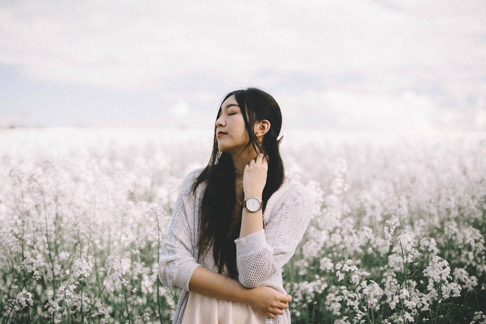 mulher vestindo pêssego parte superior interna com cardigan de malha cinza em pé atrás de campo de flores branco durante a fotografia diurna