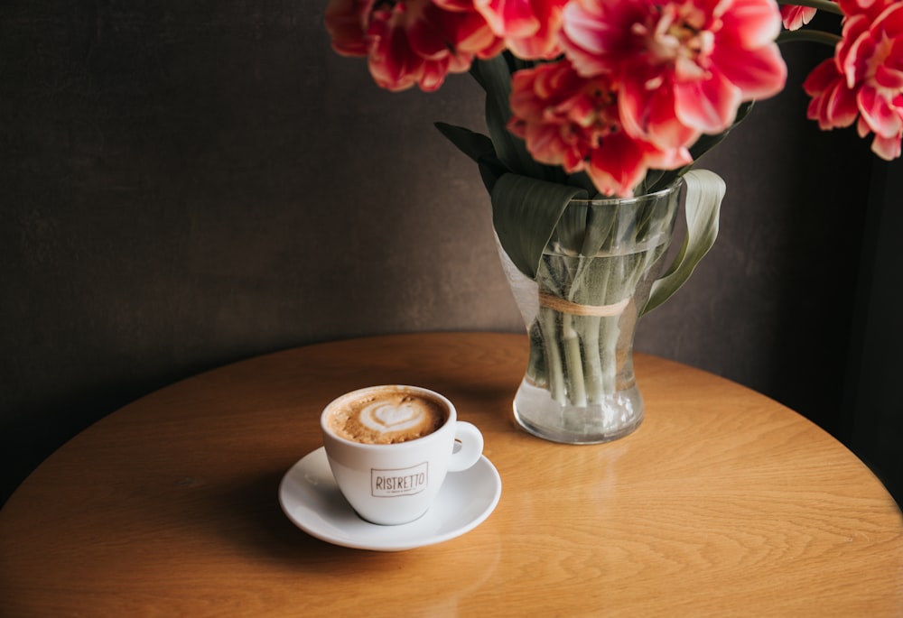 丸い茶色の木製のテーブルの上にコーヒーで満たされたカップの近くに赤い花びらの花が描かれた透明なガラスの花瓶
