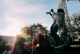 A performer is on stage energetically engaging with the crowd, while audience members cheer and raise their hands. The backlighting creates a silhouette effect around the performer. Trees and a building are visible in the background, with sunlight streaming through the scene.